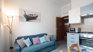 Un soggiorno moderno con un divano blu, un quadro raffigurante una barca, una lampada da terra, una cucina con forno a microonde e forno, e un tavolo da pranzo con sedie.
