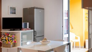 Una pequeña kitchenette con refrigerador, microondas y televisor sobre una repisa. Hay una mesa de comedor con sillas y una cesta de flores.
