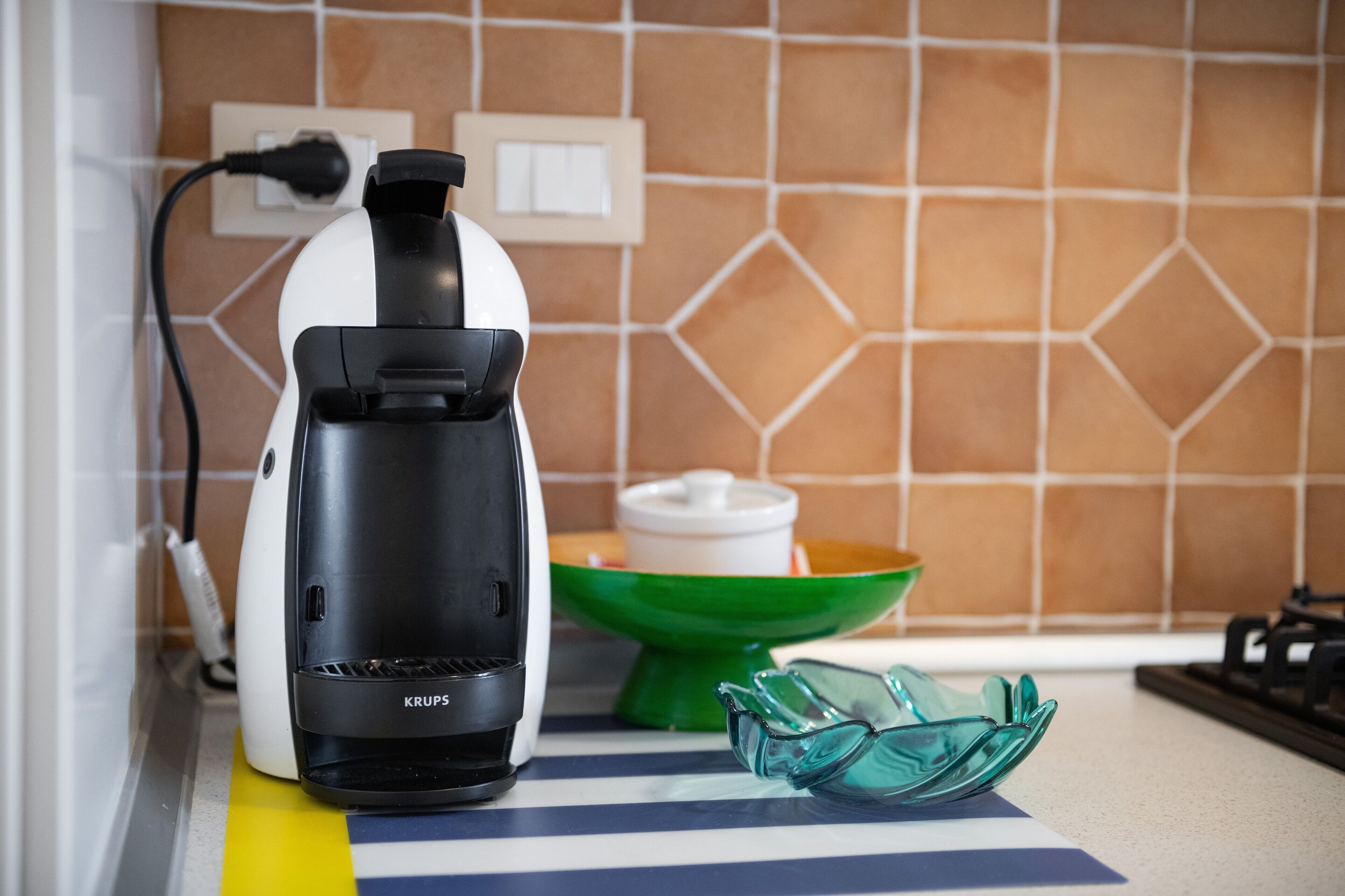 Una cocina con cafetera, un cuenco verde y un par de vasos sobre un felpudo a rayas.
