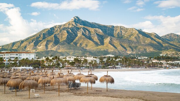 Ein Strand mit Reetdachschirmen, ein Berg im Hintergrund und Gebäude entlang der Küste.