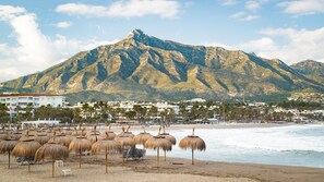 A beach with thatched umbrellas, a mountain in the background, and buildings along the shore.