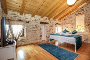 Un dormitorio con pared de piedra, vigas de madera en el techo, una cama con colcha verde, una alfombra azul y una mesita de noche con lámpara.