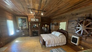 A wooden cabin room with a bed, a small table, a chair, a fireplace, and a large wooden wheel. - Rustic and Romantic Farm House (Louisa)