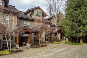 A multi-story residential building with a brick driveway, wooden railings, and a large evergreen tree.