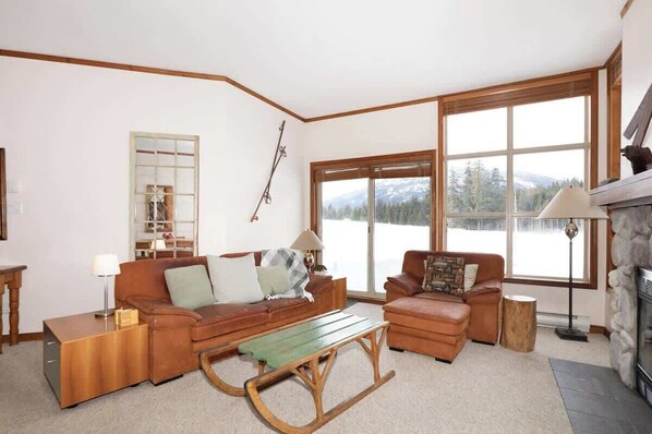 A cozy living room with a fireplace, large windows with a mountain view, and a wooden coffee table.