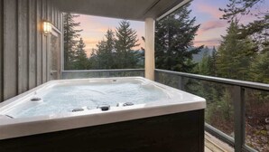 A hot tub on a wooden deck with a glass railing, overlooking a forested area.