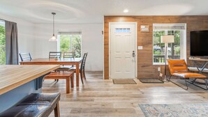 A modern living room with a wooden accent wall, a dining area with a wooden table and chairs, a TV, and a comfortable seating area with a sofa and armchair. - Walk to Downtown Sisters - DOG Friendly - Updated! (Sisters)
