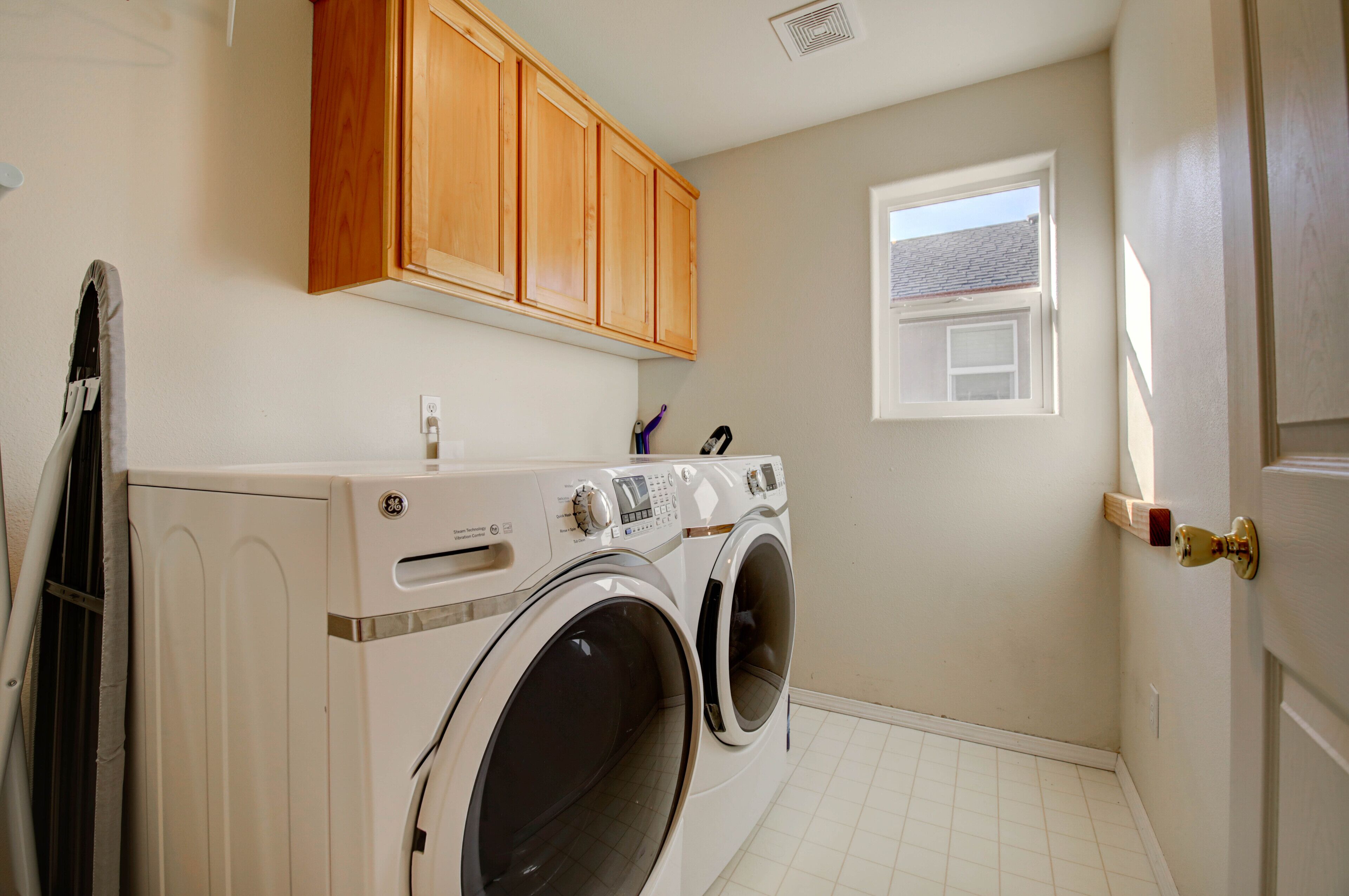 A laundry room with two front-loading washing machines, wooden cabinets above, a window, and a door with a brass handle.