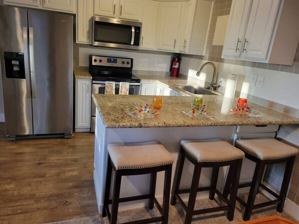 A kitchen with a stainless steel refrigerator, a microwave, a stove, and a sink. There are three bar stools and a granite countertop.