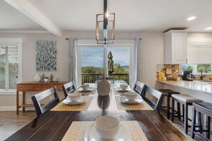 Un comedor con una mesa larga dispuesta para seis personas, una cocina con armarios blancos y un balcón con vistas.