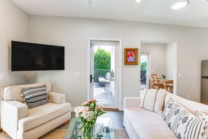 Ein Wohnzimmer mit einer Couch, einem Couchtisch mit Blumenvase, einem an der Wand montierten Flachbildfernseher und einer Tür zu einem Außenbereich.