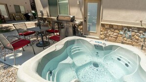 Un jacuzzi en un patio trasero con una parrilla, mobiliario exterior y un muro de piedra.