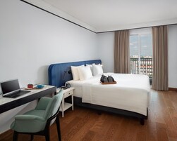 Habitación de hotel con cama, escritorio con una computadora portátil, una silla y vistas a la ciudad a través de grandes ventanales.