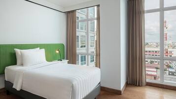 Hotellihuone, jossa on suuri sänky, vihreä pääty, yöpöytä ja suuret ikkunat, joista avautuu näkymä kaupungin siluettiin.