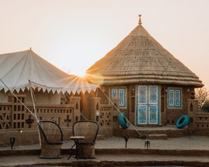 A traditional thatched-roof building with a decorative door, a white tent in the background, and two wicker chairs in the foreground. - Royal Golden Camp (Jaisalmer)