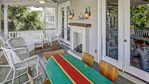 A covered porch with wicker chairs, a colorful table, a fireplace, and a view of greenery. - North Beach Tybee Home With Roof Top Deck | Walk to Beach | Heated pool access (Tybee Island)