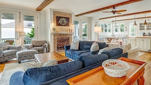 A spacious living room with a fireplace, blue sofas, and a dining area with a kitchen in the background. - North Beach Tybee Home With Roof Top Deck | Walk to Beach | Heated pool access (Tybee Island)