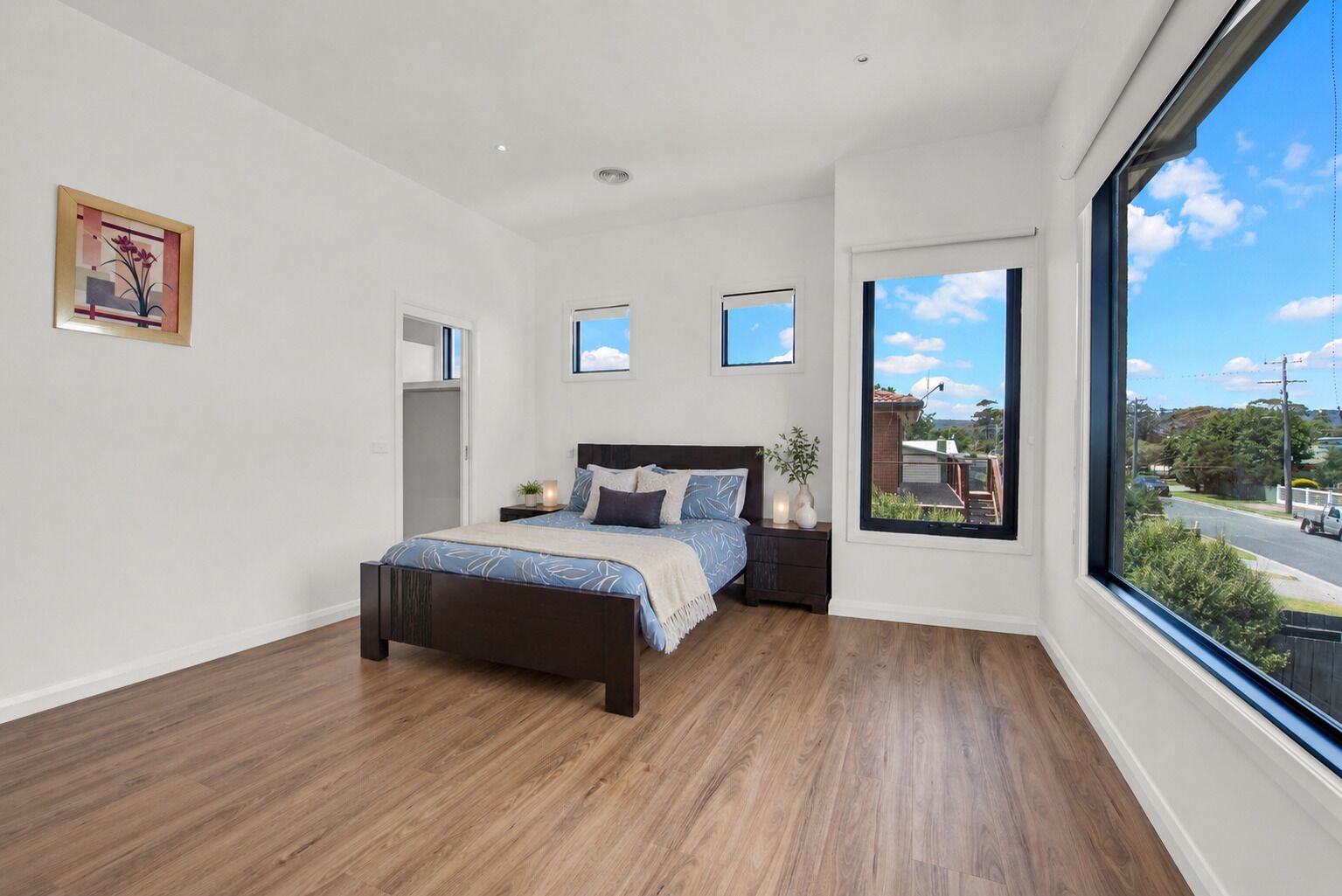 A bedroom with a large window, a bed, a nightstand, and a framed picture on the wall.