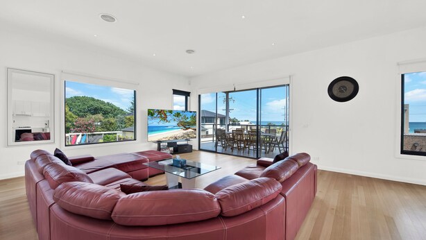 A spacious living room with a large red leather sofa, a glass coffee table, and a view of a beach through large windows. - Beach View Escape - Close to the Beach & WiFi (Safety Beach)