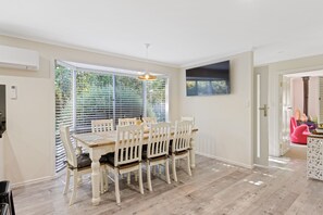 A dining area with a wooden table and white chairs, large windows with blinds, and a ceiling fan. - Bethel by the Sea by Ready Set Stay - Close to the beach & WiFi (Surf Beach)