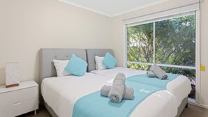Een slaapkamer met een groot raam, een bed met een grijze hoofdeinde en een nachtkastje met een lamp.