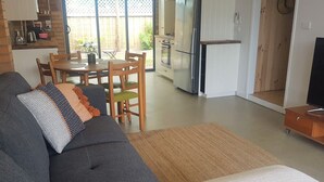 Ein modernes Wohnzimmer mit grauem Sofa, einem Esstisch mit Stühlen, einer Küchenzeile mit Kühlschrank und Backofen sowie einem Fernsehschrank.