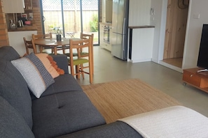 Ein modernes Wohnzimmer mit grauem Sofa, einem Esstisch mit Stühlen, einer Küchenzeile mit Kühlschrank und Backofen sowie einem Fernsehschrank.