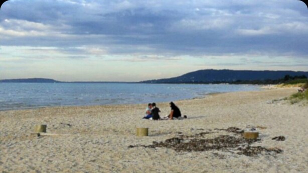 Ein Strand mit zwei Personen, die im Sand sitzen, ein paar Heuballen und ein ruhiges Meer im Hintergrund.