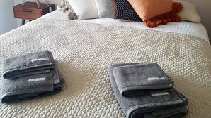 Ein ordentlich bezogenes Bett mit weißer Struktur, ein Nachttisch mit Lampe und ein grauer, gefalteter Blazer auf dem Bett.