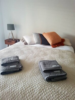 Ein ordentlich bezogenes Bett mit weißer Struktur, ein Nachttisch mit Lampe und ein grauer, gefalteter Blazer auf dem Bett.