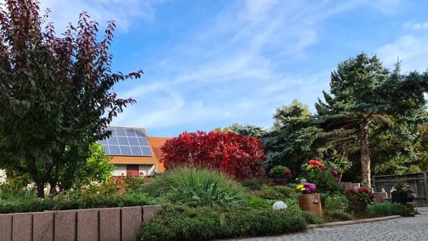 Ein gepflasterter Weg führt zu einem Gebäude mit einer Solaranlage auf dem Dach, umgeben von üppigem Grün und blühenden Pflanzen.