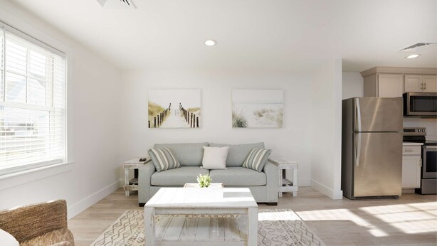 A modern living room with a grey sofa, white coffee table, and patterned rugs. There are two wall-mounted pictures, a kitchen area with a stainless steel refrigerator, and a window with blinds.