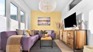 A modern living room with a purple sofa, a wooden coffee table, and a TV on a wooden cabinet. - Wolke 7 - Infrared Sauna, Fireplace, Rooftop Terrace, Bathtub, Parking Space (Olpenitz)