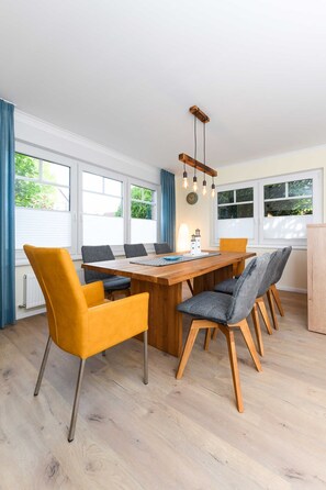 A dining area with a wooden table, six chairs, and a hanging light fixture. - Ferienwohnung Ankerplatz - Haus Meerzeit (Esens)