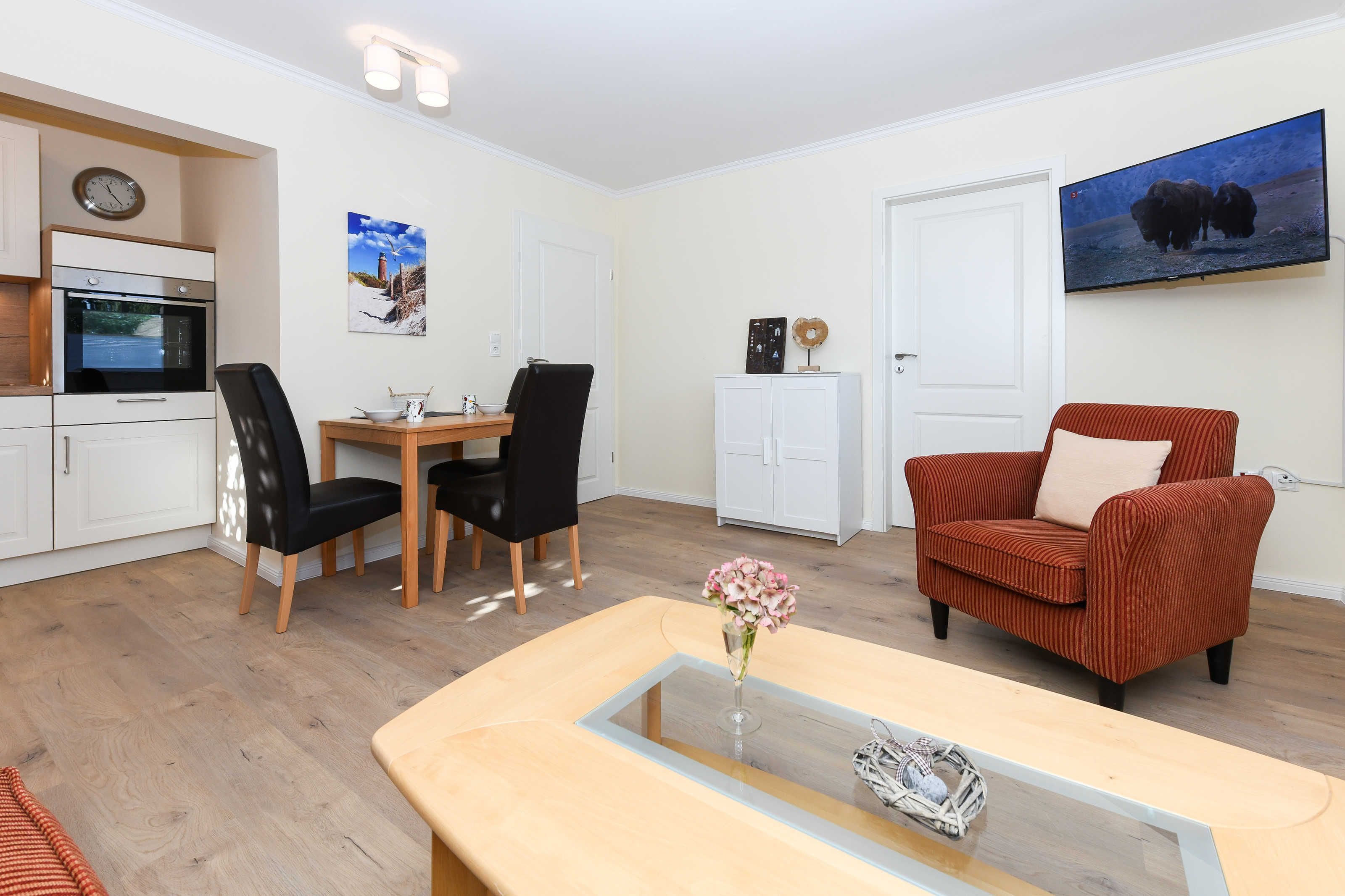 A modern living room with a dining area, a kitchen with white cabinets, a flat-screen TV, and a clock on the wall.