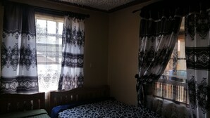 A room with two beds, one with a patterned bedspread and the other with a plain one. There are curtains with intricate designs on the windows. - smiles home (Thika)