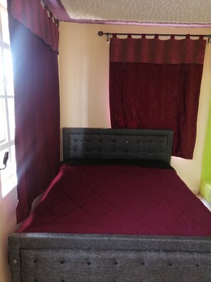 Een bed met een donker hoofdeinde, een rode sprei en een raam met paarse gordijnen.