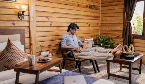 A wooden cabin interior with a person reading a book, a bed, a table with a plate of food, and a window with curtains. - Panarpani Retreat Pachmarhi (Pipariya)