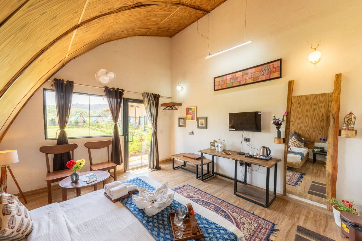 Une pièce spacieuse avec un grand lit, un bureau et une télévision. Il y a une fenêtre avec des rideaux et une vue sur la verdure à l’extérieur.