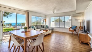 A spacious living room with a wooden dining table, a sofa, and a TV stand. Large windows offer views of the outdoors. - THE GRASSY KNOLL Panoramic Beach Apartment  - Sunrise Beach (Sunrise Beach)