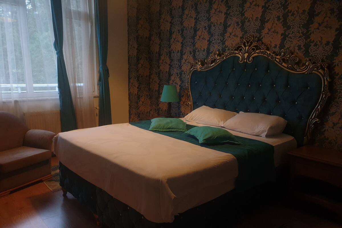 A neatly made bed with a green bedspread and white pillows, a wooden nightstand with a lamp, and a chair in a room with patterned wallpaper and a window with sheer curtains.