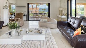 A spacious living room with a large window, a sofa, a coffee table, and a dining area visible through the glass doors. - Villa at the entrance to St Tropez sea view beach 2 minutes walk (Gassin)