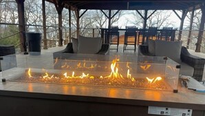 Un foyer au gaz avec un plateau en verre, installé dans un espace extérieur abrité, agrémenté de meubles en osier et offrant une vue sur les arbres.