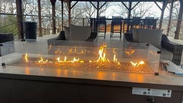 A glass-top fire feature in a covered outdoor seating area with wicker furniture and a view of trees.
