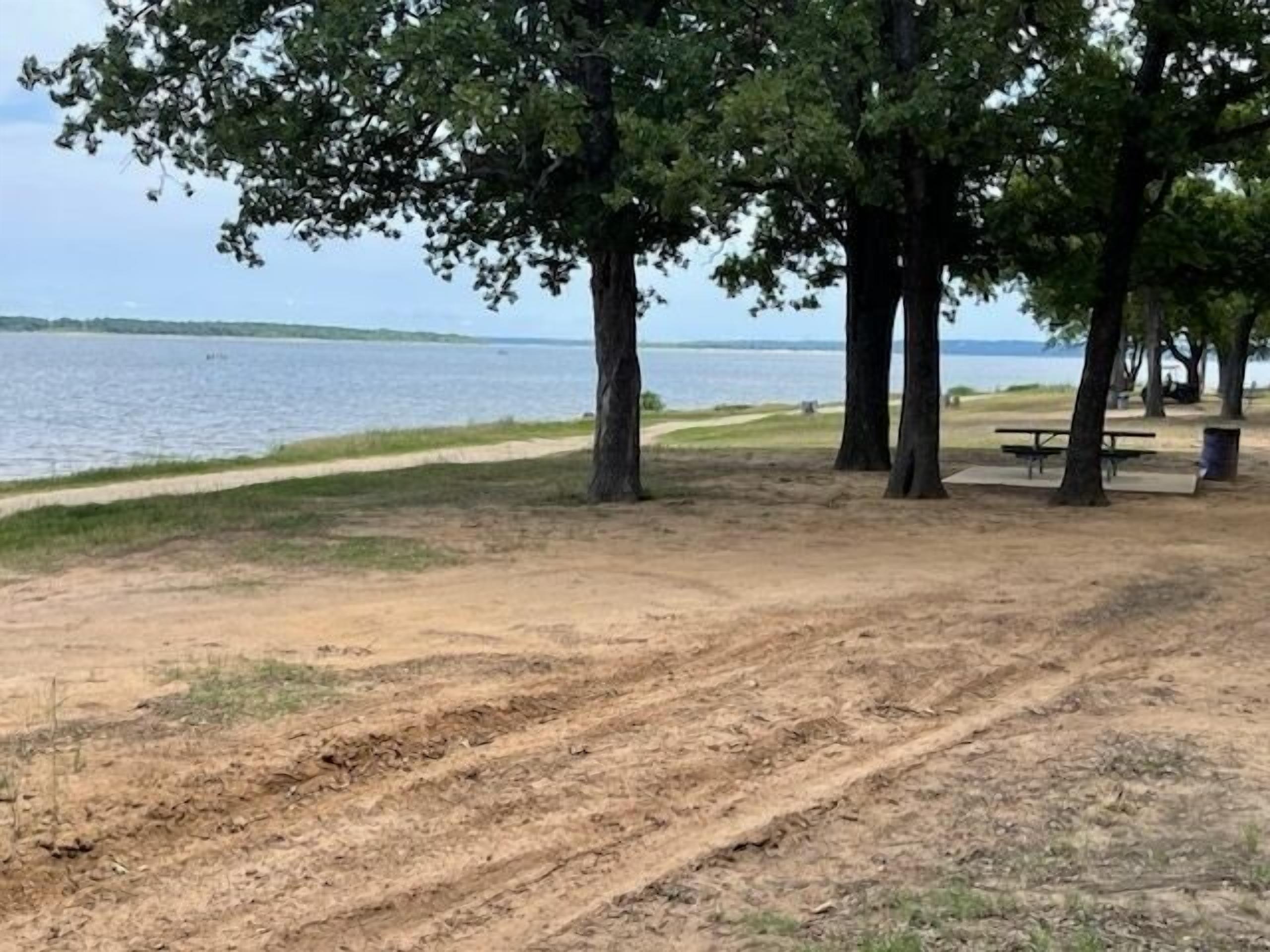 Un parc avec un sentier de terre, des tables de pique-nique et des arbres, situé près d’une étendue d’eau.