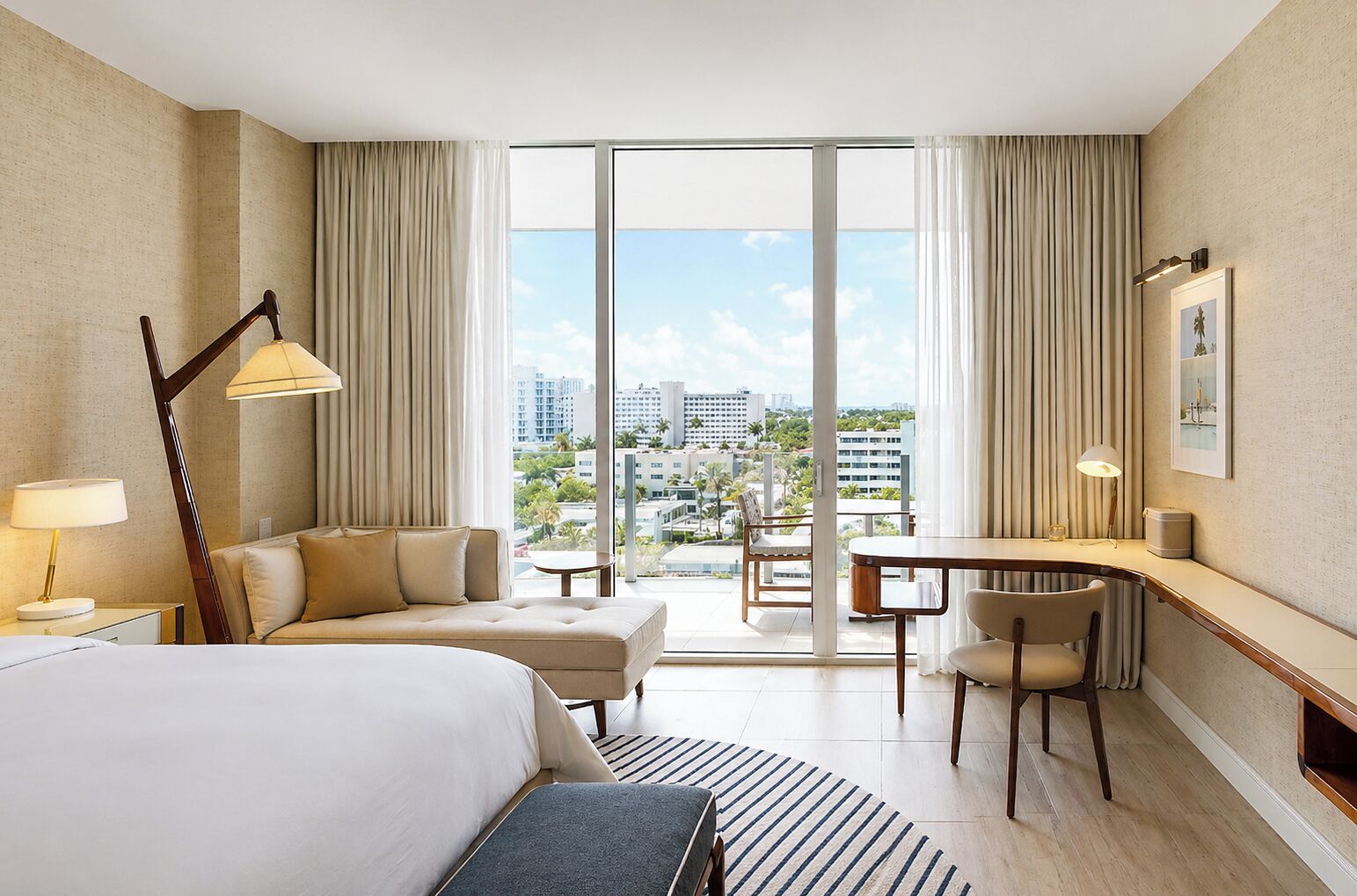 A hotel room with a bed, a desk, a chair, a sofa, a lamp, a picture on the wall, and a view of the city through large windows.