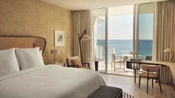 Quarto de hotel com uma cama grande, uma escrivaninha com cadeira, uma mesinha e vista para o oceano.
