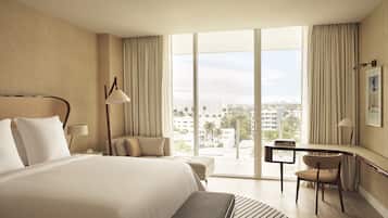 A hotel room with a large bed, a desk, a chair, and a view of the city through large windows.