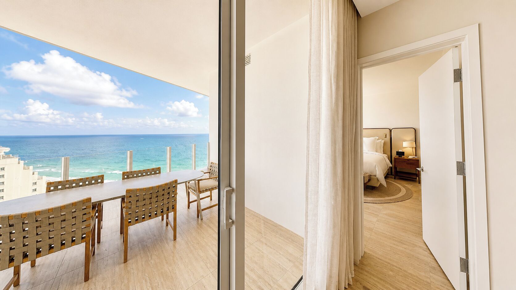 A balcony with a view of the ocean and cityscape, a dining area with a table and chairs, and a bedroom with a bed and nightstand.