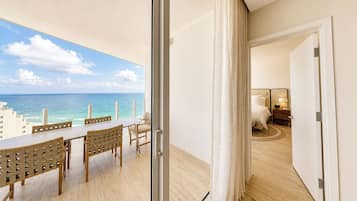 A balcony with a view of the ocean and cityscape, a dining area with a table and chairs, and a bedroom with a bed and nightstand.